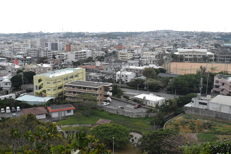 安慶名城跡から安慶名1～3丁目方面を望む。平らな土地が広がり、中層の建物が多い。写真右手には見えるのは、あげな中学校のグラウンド