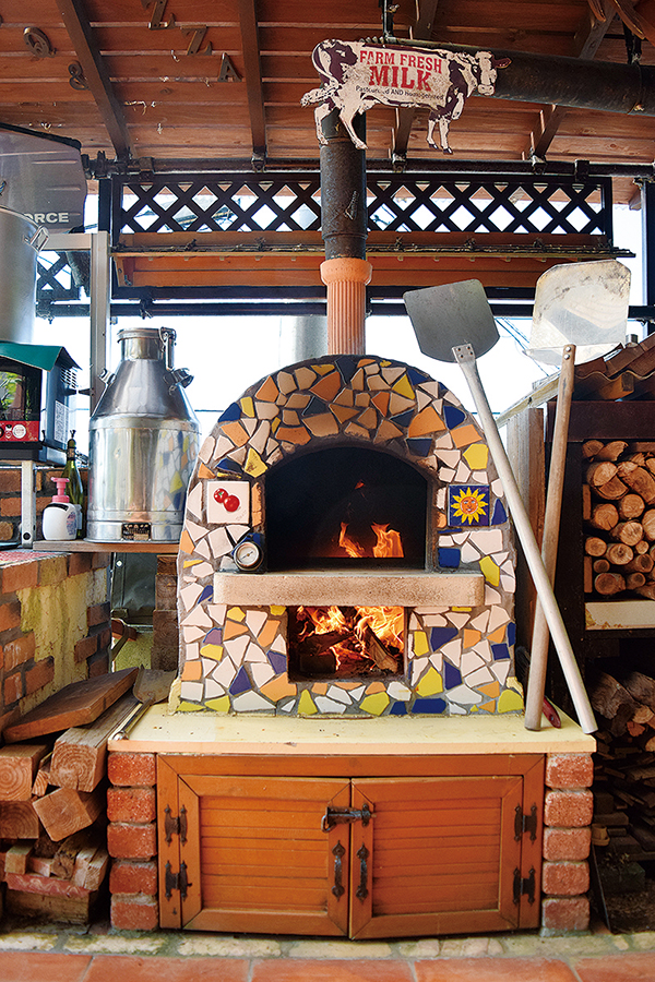 ２層式のピザ窯は奥で上下がつながっている。短かった煙突は、ダクトを曲げてパーゴラの上まで延ばしたことで火力がアップ。ミルク缶（左）などは母親の好みで集めているものだそう