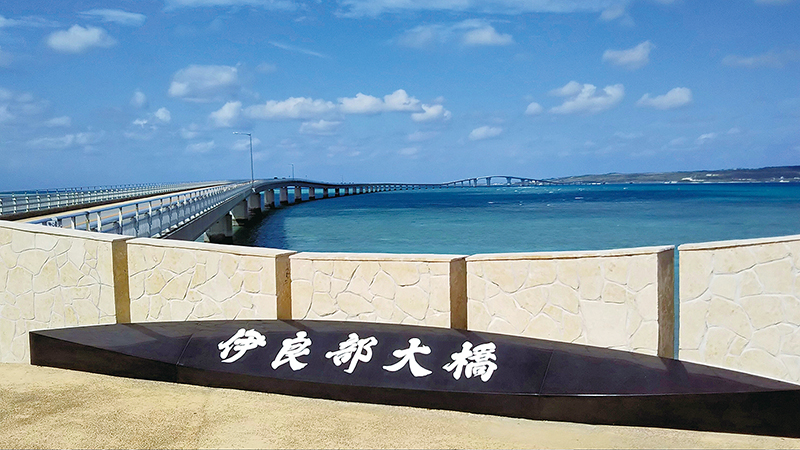 橋名の看板の後ろに延びるのが、全長３５４０㍍、通行料金を徴収しない橋としては日本一長いと言われる伊良部大橋