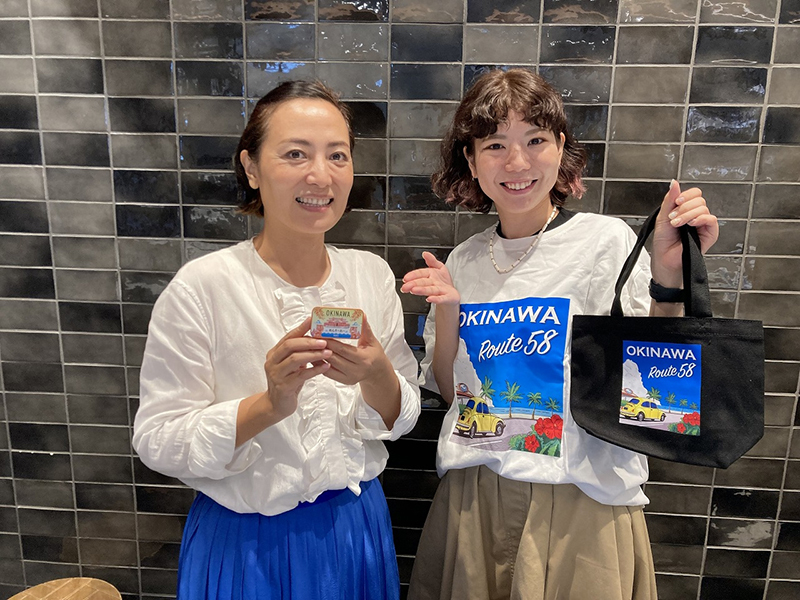 那覇の事務所で商品をアピールする、ブルーリンクの大坂美恵子さん（左）と、ミント缶のファンと称する湧川日奈子さん。ミント缶と同じデザインのＴシャツとバッグ（非売品）を着用